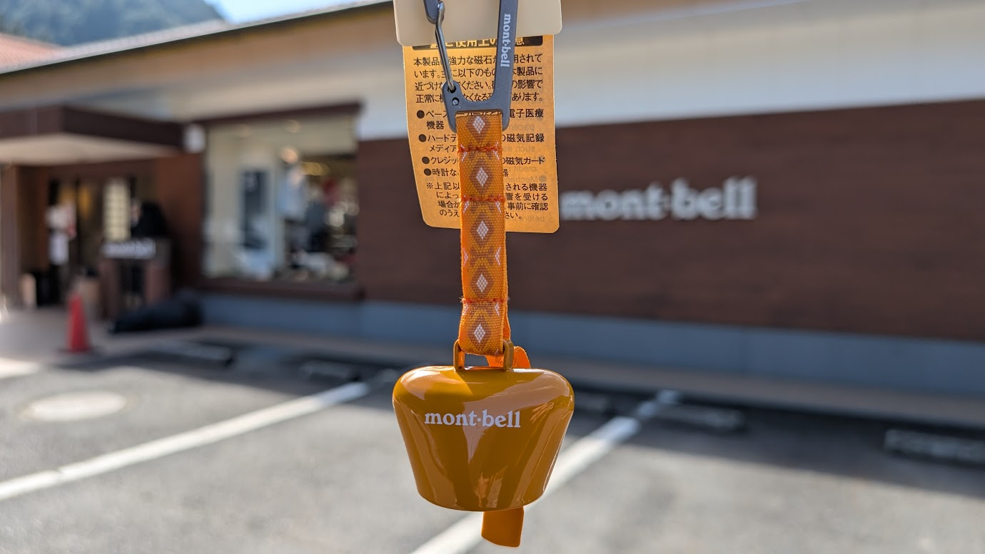 奈良県黒滝村の「道の駅 吉野路黒滝」にあるモンベルの店舗前で、購入したオレンジ色の熊鈴を手に持っている。背景にはmont-bellのロゴが見える。