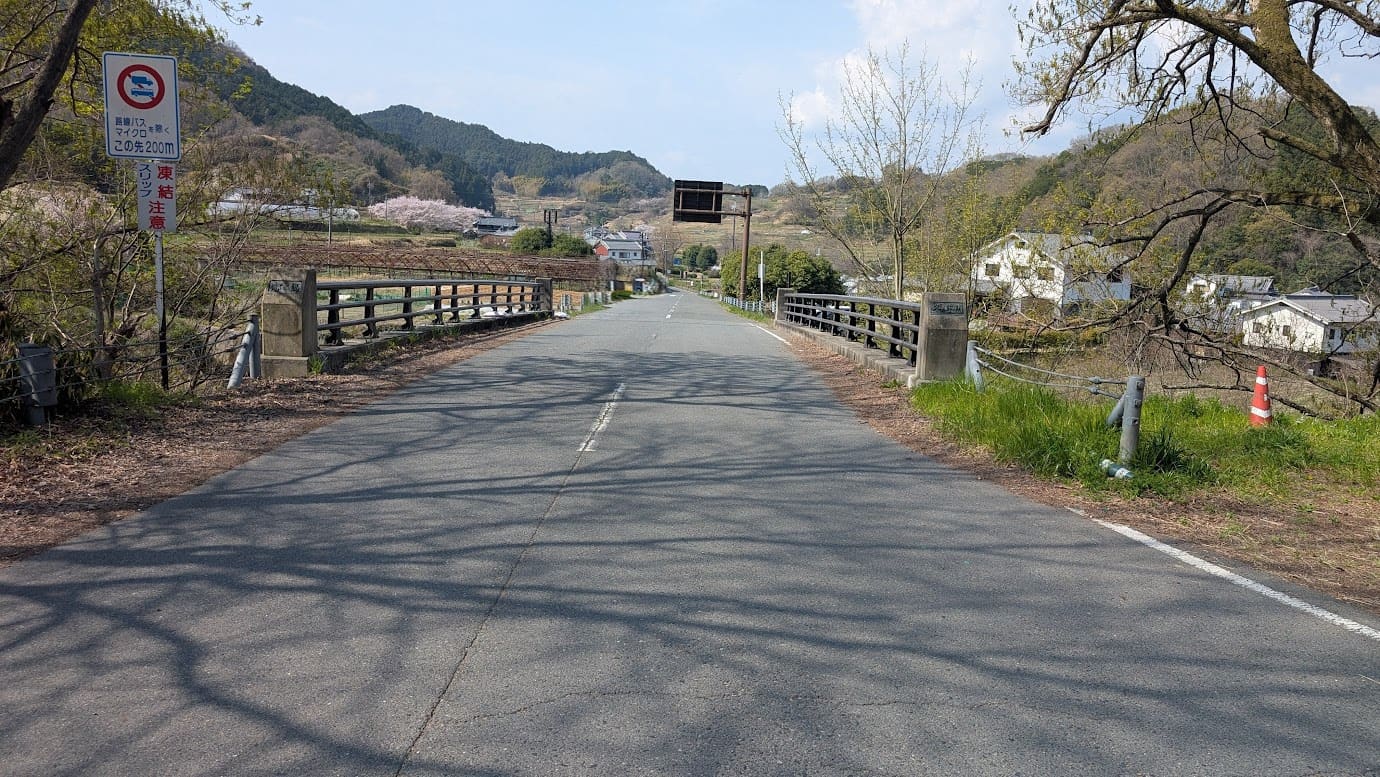 奈良県明日香村、都塚古墳へと続く道に架かる「細川橋」。アスファルトの道には木の影が落ち、前方には飛鳥の古い町並みとのどかな山並みが広がっている風景(2026年3月30日撮影)。