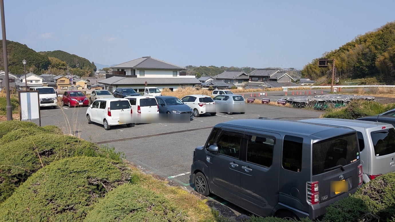 奈良県明日香村、石舞台古墳の無料駐車場内部。平日の昼間（2026年3月30日撮影）にもかかわらず、15台分のスペースはすべて埋まっており、軽自動車や普通車がぎっしりと駐車されている。背景には明日香村の町並みが見える風景。