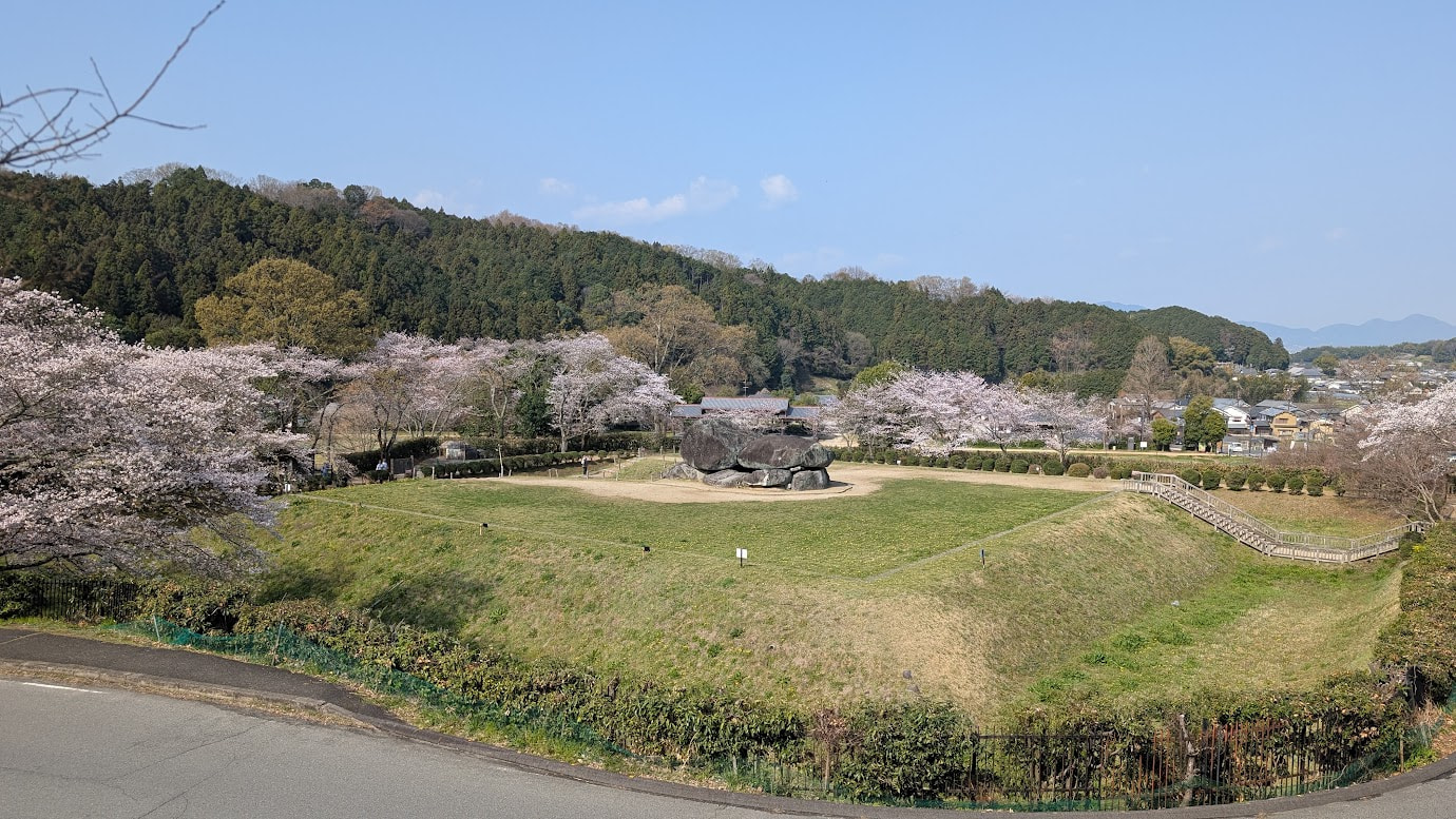 奈良県明日香村の石舞台古墳の全景。高台からの視点で、中央に露出した巨石の石室があり、その周囲を広い芝生と満開の桜並木が囲んでいる。遠くには飛鳥の山々と民家が見える春の風景（2026年3月30日撮影）。