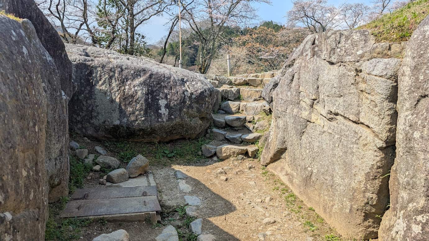 石舞台古墳の入り口付近にある、幅が細くなった石の階段。一人が通るのがやっとの幅で、両脇は芝生と石垣に囲まれている（2026年3月30日撮影）。