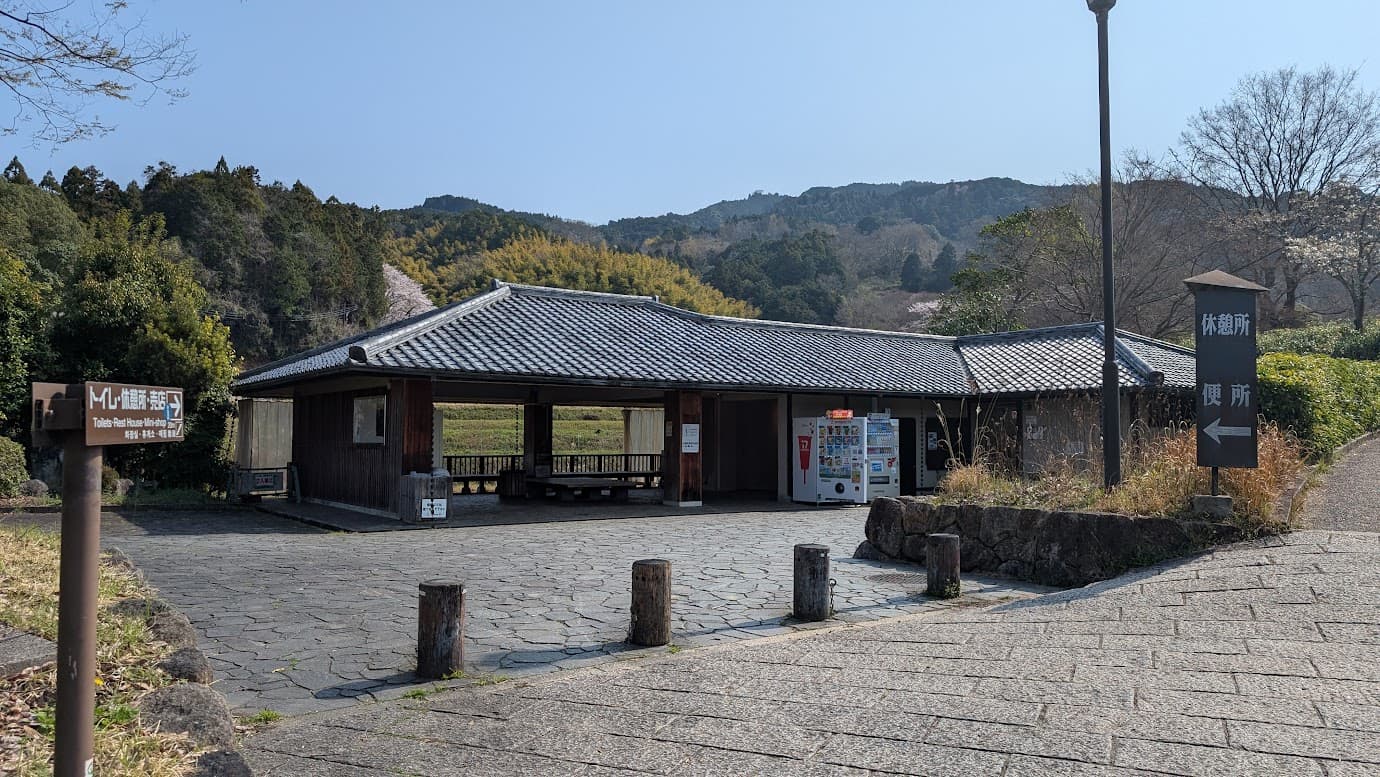 奈良県明日香村、石舞台古墳のすぐ近くにある瓦屋根の休憩所と公衆トイレ。石畳の広場に面しており、建物内にはベンチや自動販売機が設置されている。「便所」「休憩所」と書かれた木製の案内看板が立つ風景（2026年3月30日撮影）。