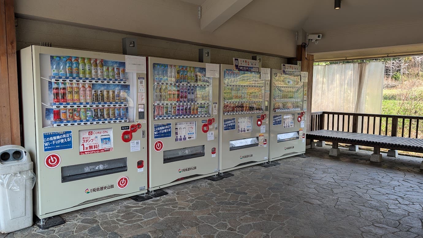 奈良県明日香村の石舞台古墳前にある休憩所内部。飲料の自動販売機が4台並んで設置されており、電子マネー対応やWi-Fiスポットの案内も見える。横には木製のベンチがあり、散策の合間に休憩できるスペース。