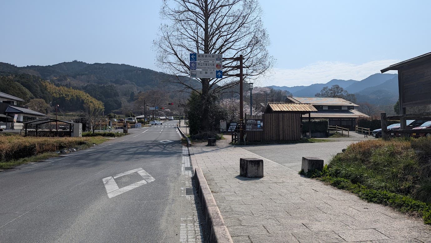 奈良県明日香村の石舞台古墳へと続く道路。青空の下、「石舞台 ↑」「飛鳥資料館 ←」などの案内看板が立つ分岐点。歩道は石畳で整備されており、周囲には飛鳥の山々と民家が広がる風景（2026年3月30日撮影）。