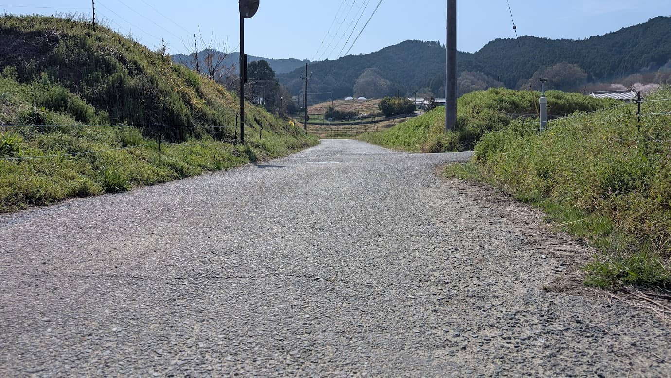 奈良県明日香村、都塚古墳のすぐ手前にある緩やかな上り坂の舗装路。周囲は低い緑の丘に囲まれており、青空に向かって道が伸びている。右側には電柱が立ち、遠くには飛鳥の山並みが広がる風景(2026年3月30日撮影)。