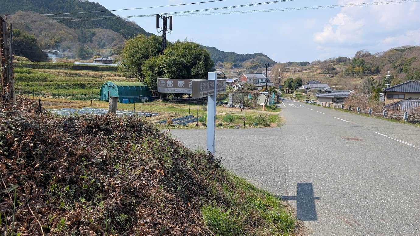 奈良県明日香村、都塚古墳へと続くT字路の案内標識。「← 都塚古墳 0.1km」と書かれた看板があり、前方の道は緩やかに下りながら集落へと続いている。周囲にはビニールハウスや飛鳥の山々が広がる風景(2026年3月30日撮影)。