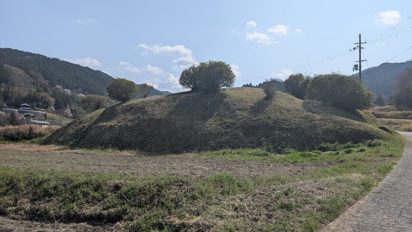奈良県明日香村にある都塚古墳の全景。斜面が階段状に石積みされたピラミッドのような特異な構造をしており、周囲は緑の芝生と木々に囲まれている。青空の下に静かに佇む歴史的遺構(2026年3月30日撮影)。