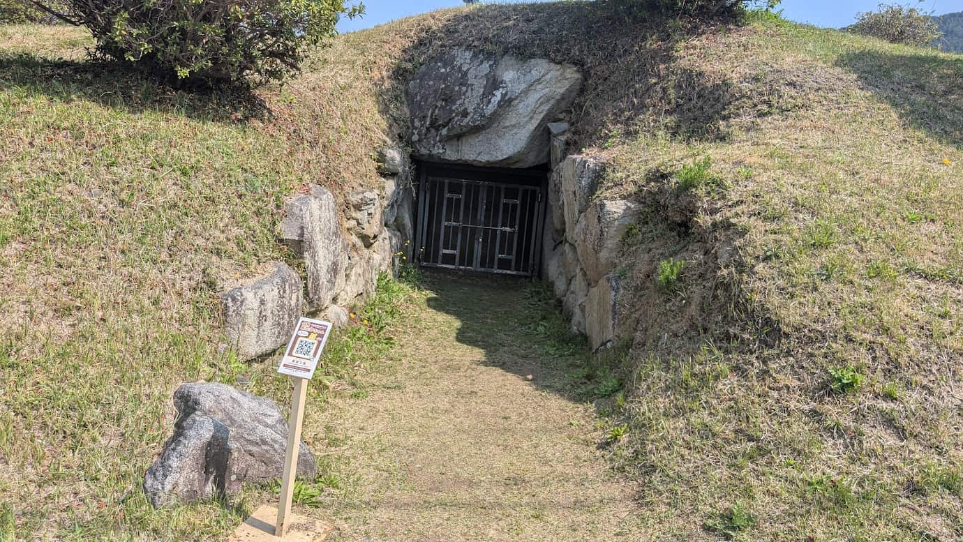 奈良県明日香村、都塚古墳の石室入口。巨大な天井石と側壁の石組みに囲まれた開口部には鉄格子の扉が設置されており、手前には解説用のQRコード看板が立っている。春の柔らかな日差しが差し込む古墳の入り口(2026年3月30日撮影)。