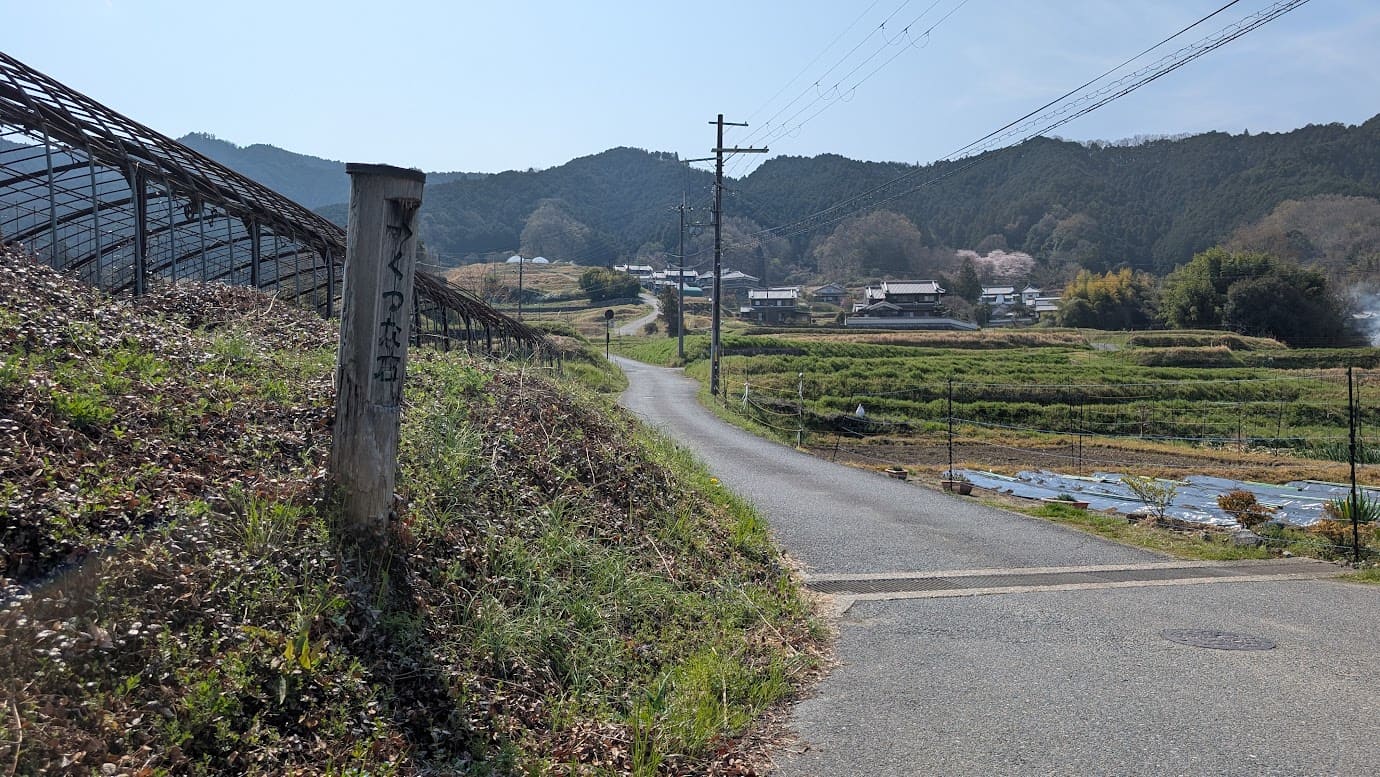 奈良県明日香村、都塚古墳の近くにある散策路。手前の古い木柱には「くつな石 →」と手書きで刻まれている。道は緩やかなカーブを描いて集落の奥へと続き、背後には飛鳥の棚田と山々が広がっている(2026年3月30日撮影)。