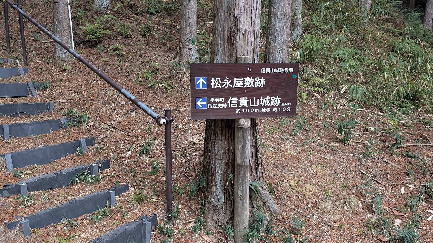 信貴山城跡の深い森の中に立つ「松永屋敷跡」と書かれた白い標柱。周囲は高く伸びる杉の木々に囲まれ、足元には落ち葉が積もっている。