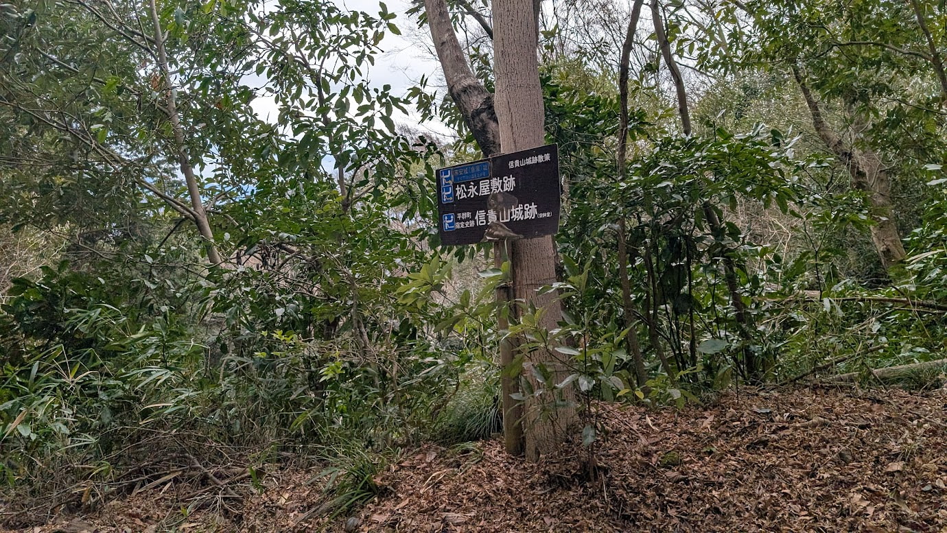 森の中の大きな木に取り付けられた「松永屋敷跡」「信貴山城跡」と書かれた黒い案内看板。周囲は深い緑の木々に囲まれており、歴史散策路の途中に位置している。