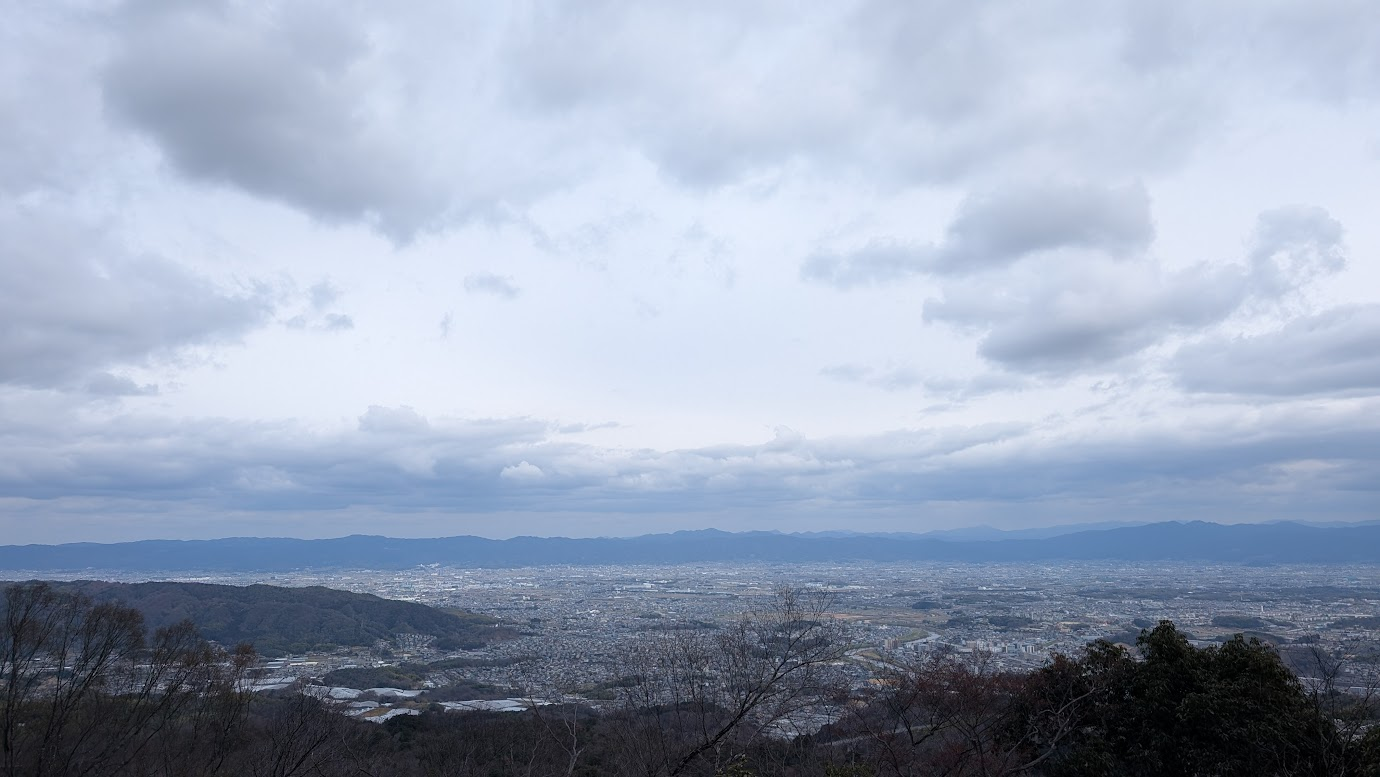 信貴山城址の遺構越しに広がる奈良盆地の全景。手前には城跡を思わせる急峻な地形や木々があり、その向こうに広大な大和平野が広がっている。