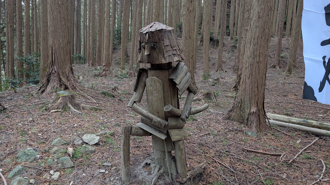 信貴山城址の深い森の中。苔むした石畳の道の脇に、真っ直ぐな杉の木を彫って作られた武士の像が立っている。武士は和装で、左手に刀を持ち、静かに佇んでいる。