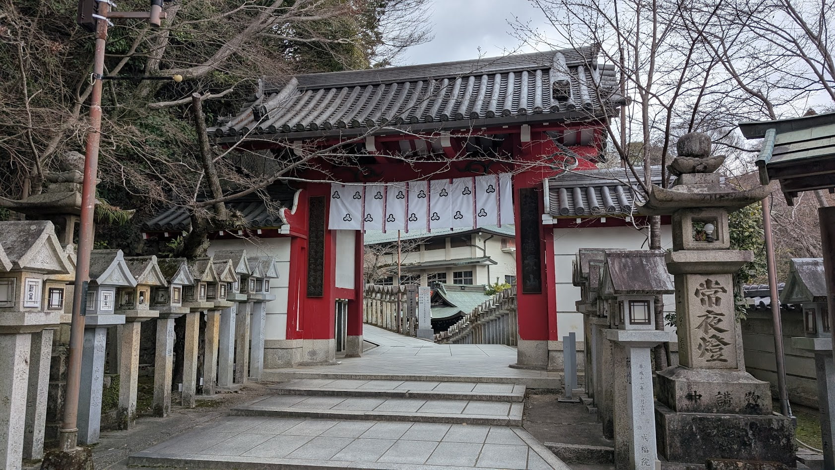 信貴山朝護孫子寺の赤門。鮮やかな朱塗りの楼門で、周囲の緑の木々とのコントラストが美しく、門の先には宿坊へと続く参道が見える。