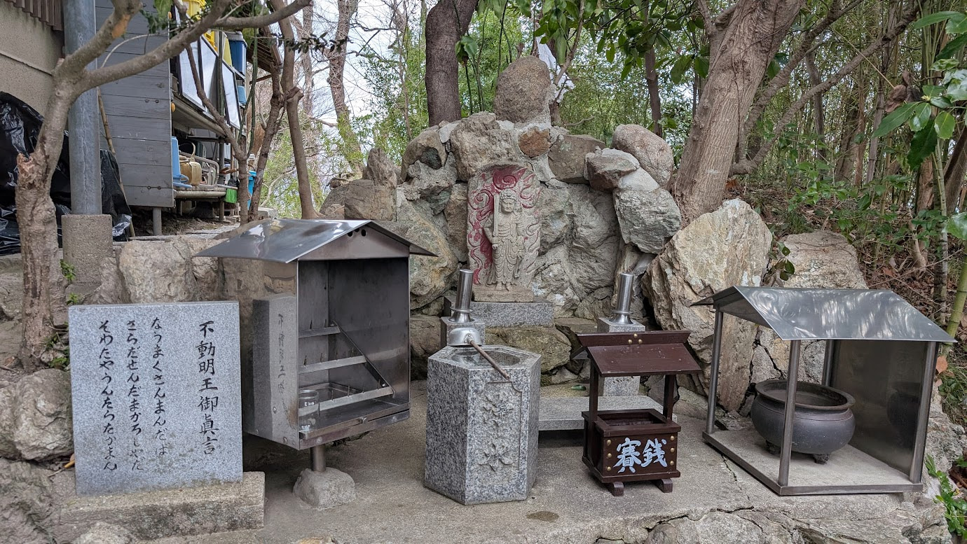 信貴山・空鉢護法堂の堂内。最奥の祭壇に、黄金の炎(迦楼羅炎)を背負った、憤怒の表情の不動明王像が鎮座している。手前には、お供え物や多くの灯籠が並び、神聖な空気が漂っている。