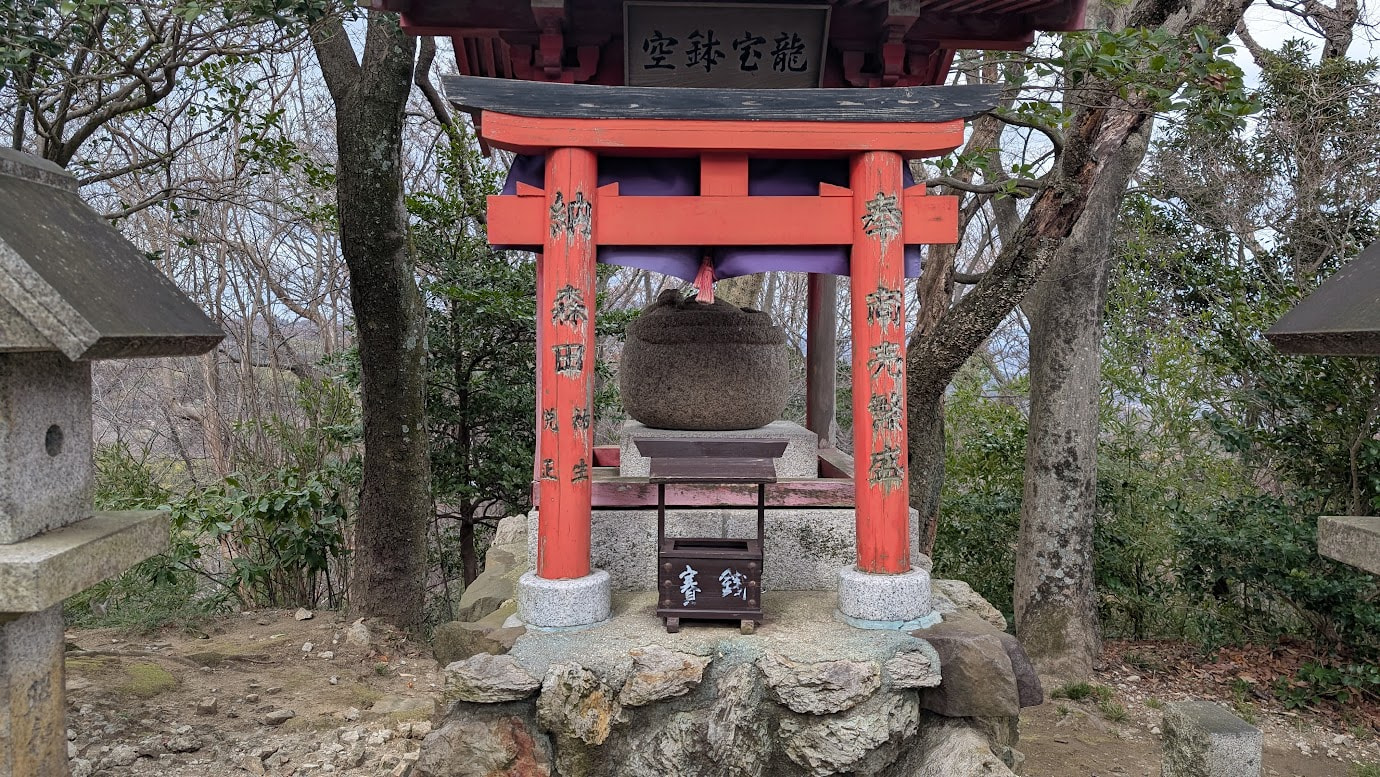 空鉢護法堂の裏手にある「空鉢宝龍」のお社。鮮やかな朱色の鳥居の中に、伝説の空鉢を象った丸い石像が祀られている。周囲は静かな森に囲まれている。