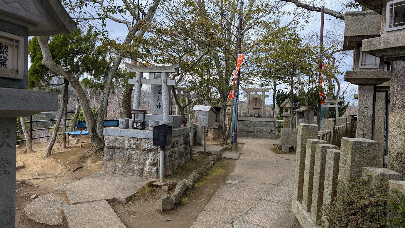 信貴山頂の空鉢護法堂。舞台にある三体の巨大な難陀龍王(蛇神)の石像の手前には、奉納された小さな巳さんの石像が並び、背景には「白瀧大神」の鳥居と石造の不動明王像、そして奈良盆地の絶景が広がっている。全体が一望できる。