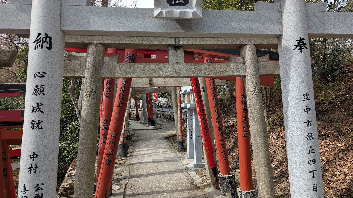 信貴山・空鉢護法堂の目前に建つ、石造りと朱色の鳥居が幾重にも重なり合う光景。鳥居には奉納者の名前が刻まれ、奥へと続く参道を包み込んでいる。