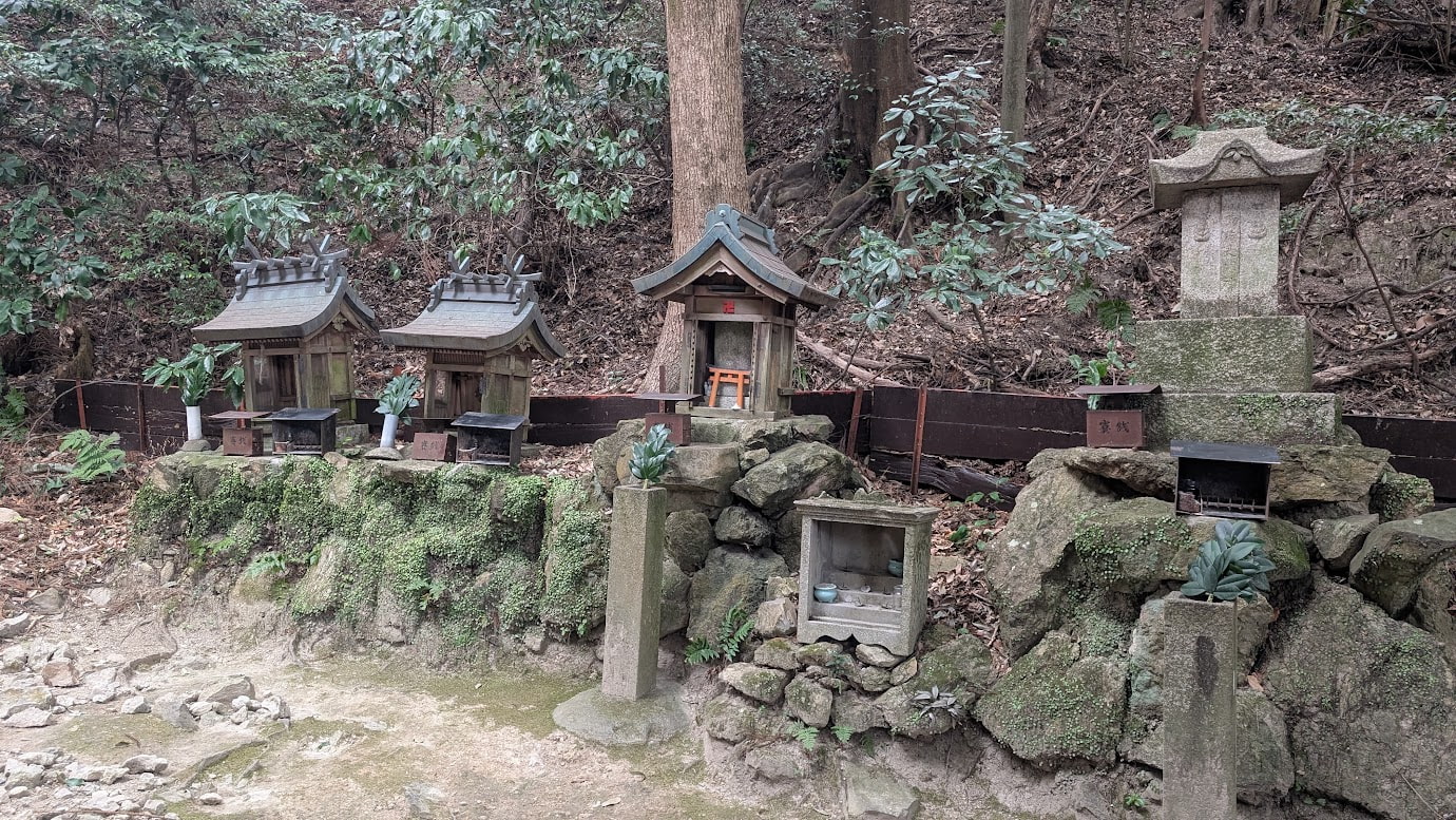 信貴山・空鉢護法堂へと続く山道の途中、苔むした岩肌に沿って並ぶ多種多様な小さな摂社・末社。木造のお社や石の祠があり、周囲は深い森に囲まれている。