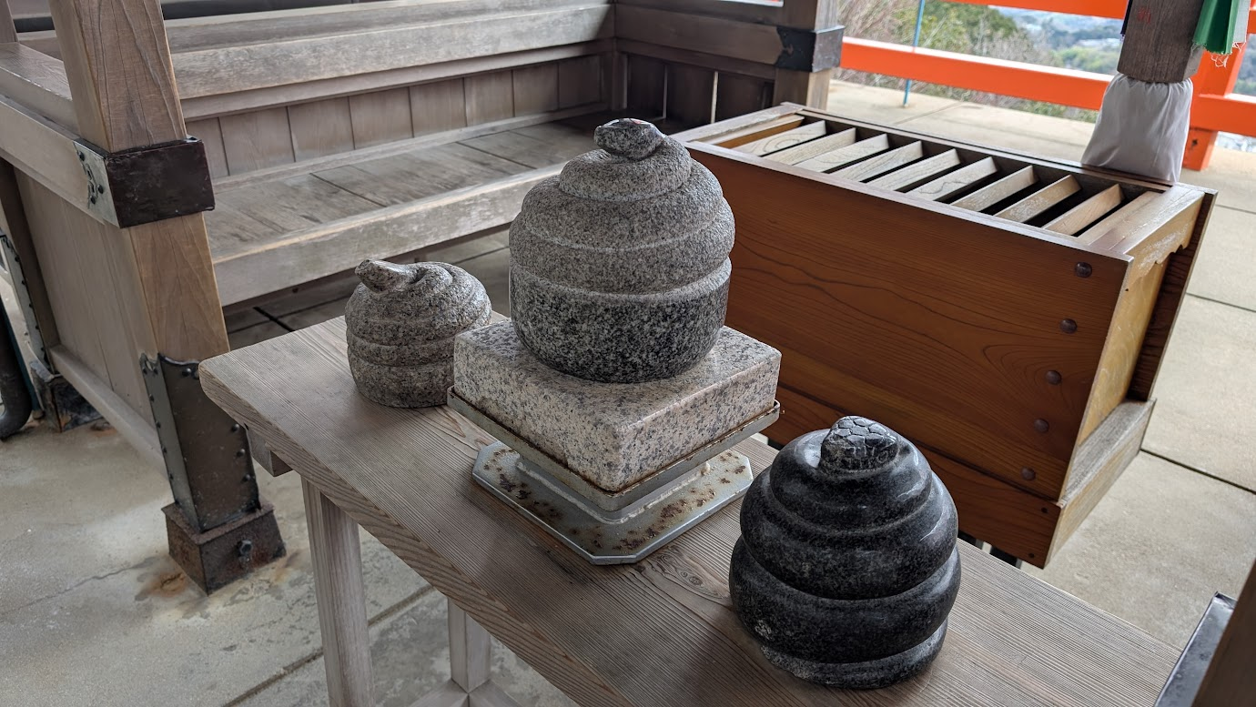 信貴山頂の空鉢護法堂舞台にある、三体の巨大な石造りの蛇神「難陀龍王」。その手前の木のベンチには、奉納された無数の小さな「巳さん」の石像が並び、背景には奈良盆地の絶景が広がっている。