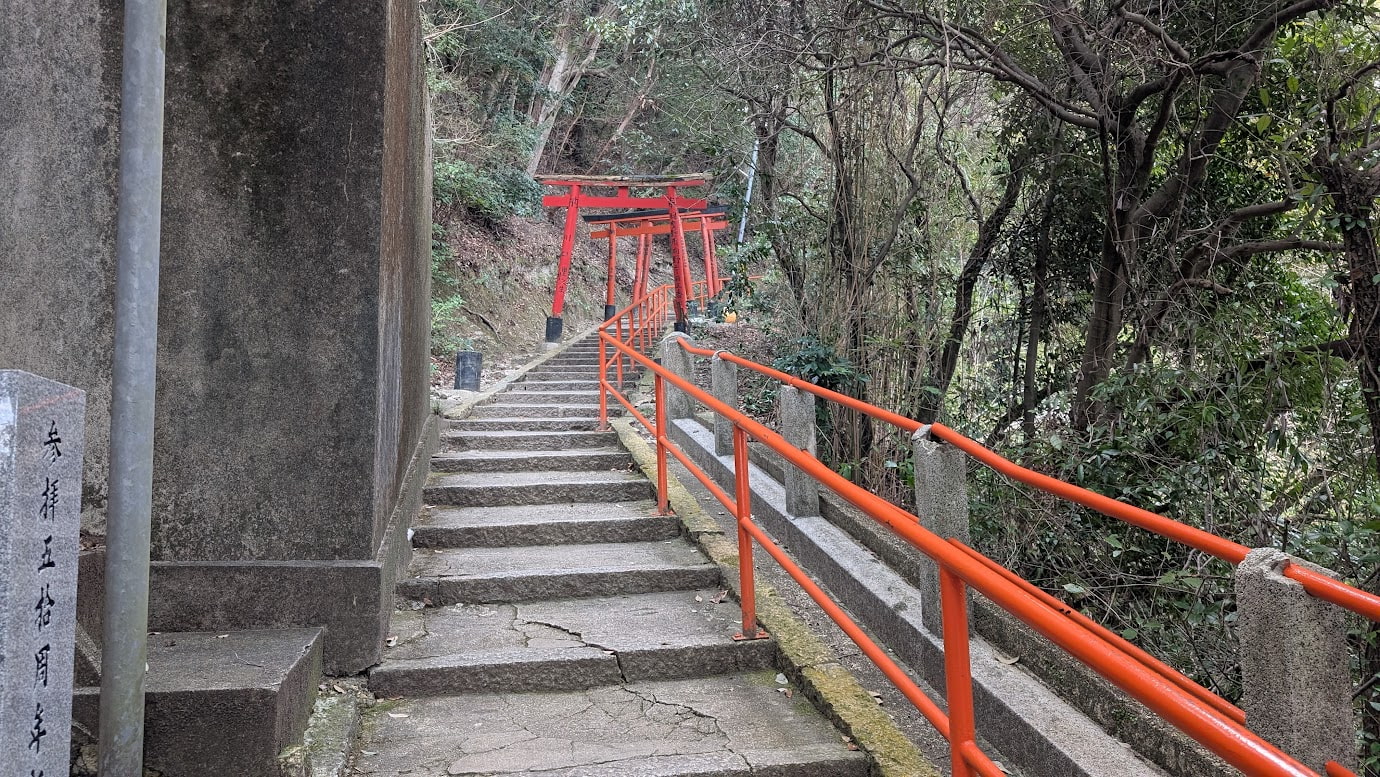 信貴山朝護孫子寺・空鉢護法堂へと続く参道の入り口。真っ直ぐに伸びる急な石段と、その上に連続して並ぶ朱色の鳥居。両脇には緑の木々が茂っている。