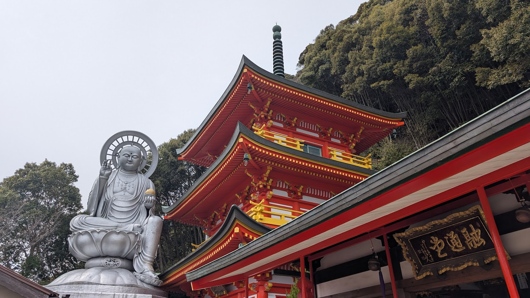 信貴山朝護孫子寺・玉蔵院にある巨大な日本一大地蔵尊(銀色)と、その横に建つ多層の鮮やかな朱色の融通堂、そして背後の森。全体が一望できる広角画像。