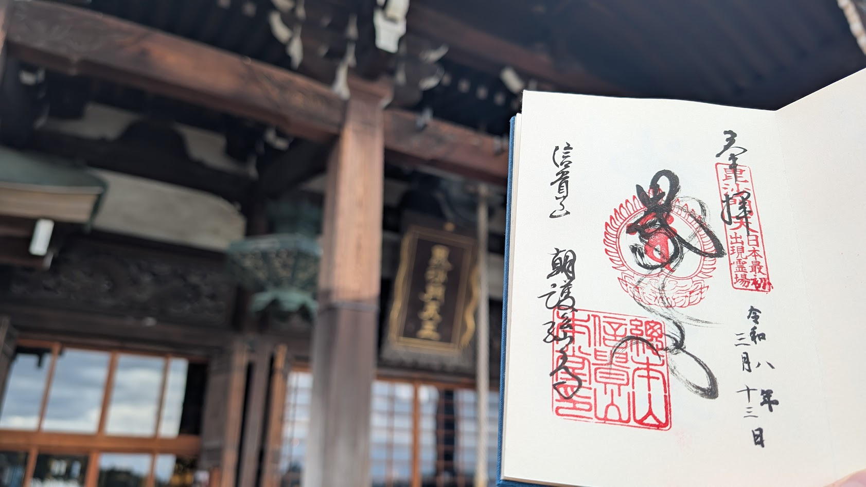 信貴山朝護孫子寺の本堂を背景に、手前に広げた御朱印帳。力強く書かれた「毘沙門天」の墨書きと朱印が、本堂の重厚な建築と重なる。