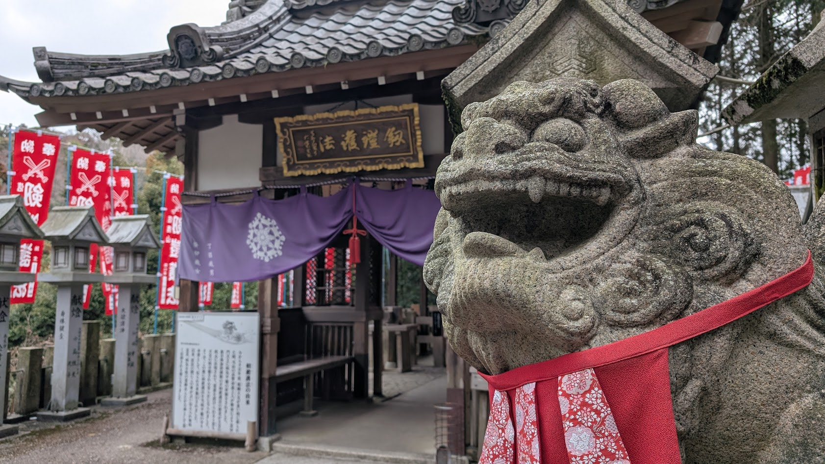 信貴山朝護孫子寺の異形の狛犬「剱鎧護法(けんがいごほう)」の阿形(あぎょう)のアップ。鎧のような皮膚と、鋭い牙、そして背後にそびえる本堂の屋根が写っている。ポートレート撮影。