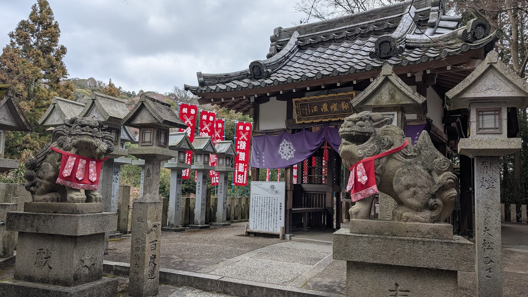 信貴山朝護孫子寺の本堂へと続く参道の脇、剱鎧護法堂の入り口を守る一対の狛犬。石造りの鳥居や石灯籠が並ぶ画像。