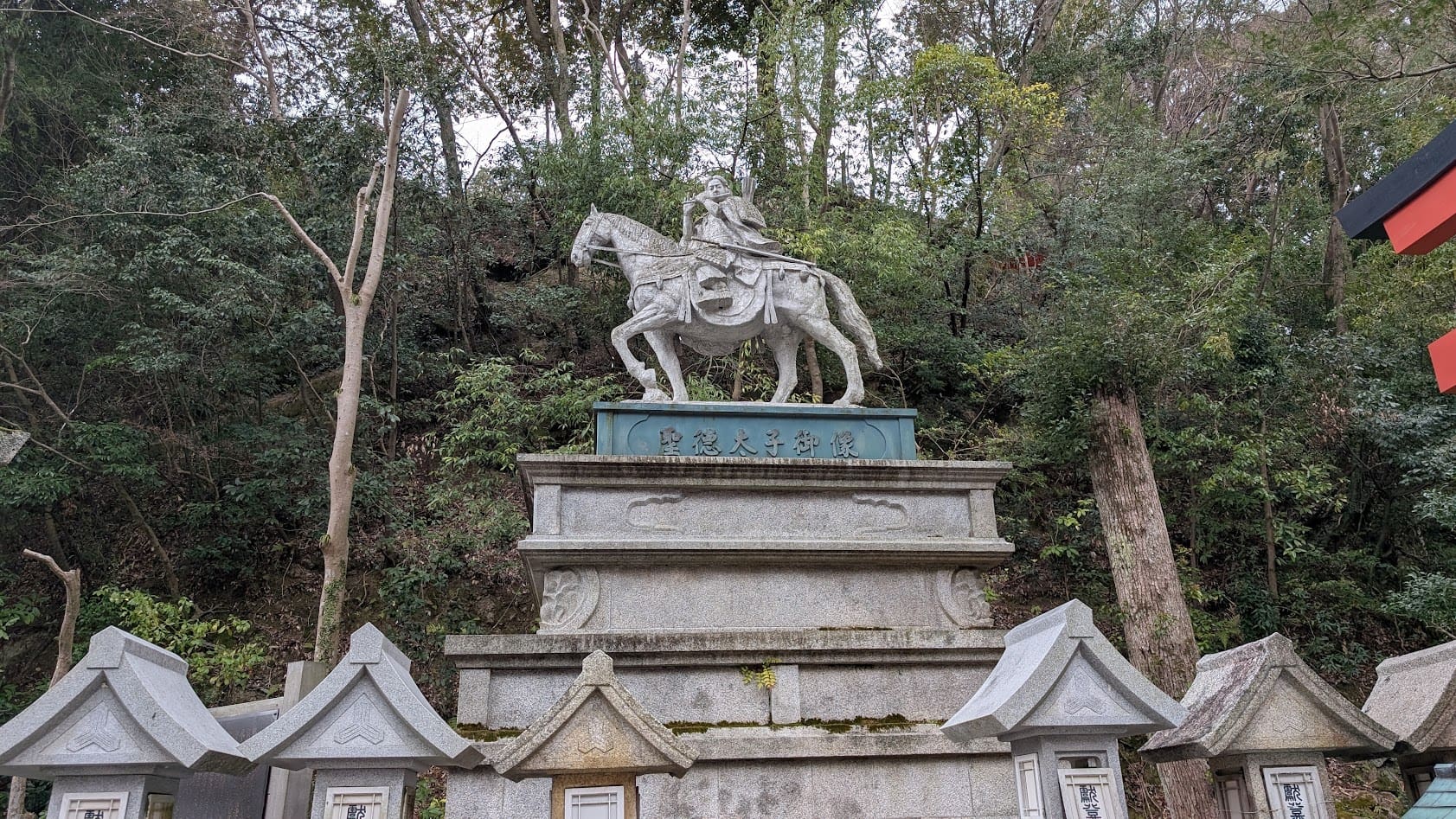 信貴山朝護孫子寺の参道に立つ、聖徳太子の立像。笏を手に、威厳を持って参道を見守る姿。周囲は緑の木々に囲まれ、石灯籠が並んでいる。