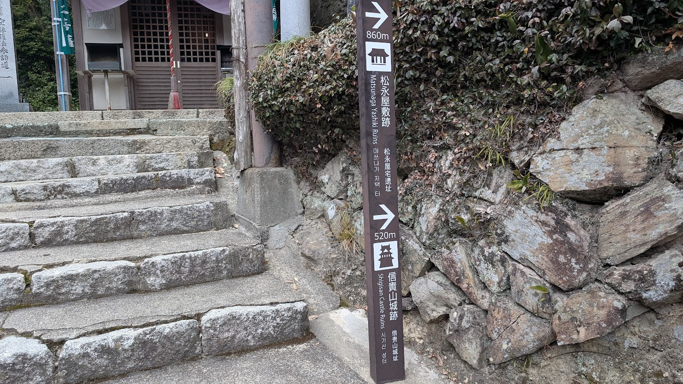 信貴山朝護孫子寺の多宝塔付近にある、石段沿いの案内標識。「←松永屋敷跡 860m」「→信貴山城址 520m」と書かれており、戦国時代の歴史スポットへの分岐点を示している。