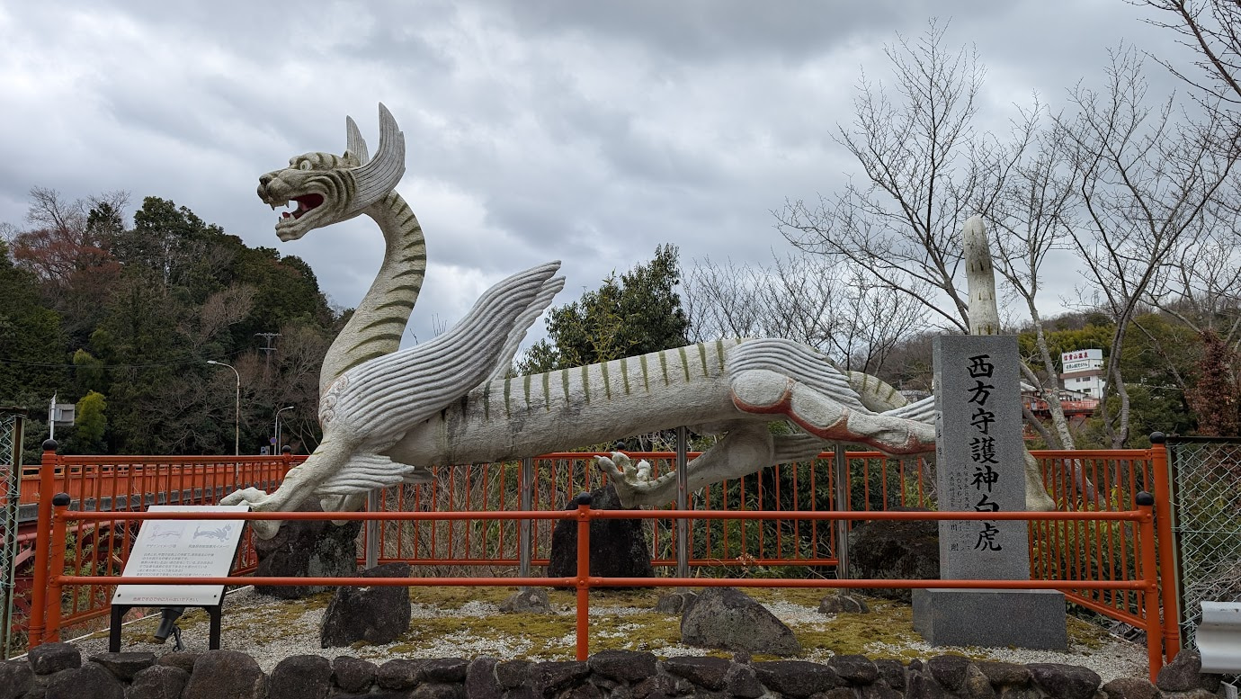 信貴山にある西方守護神「白虎」の巨大な白い石像。翼を持ち、長い体をくねらせて西を向いている。傍らには「西方守護神 白虎」と刻まれた石碑が立っている。
