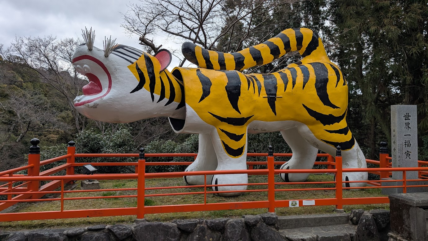 信貴山朝護孫子寺の参道に鎮座する、世界一巨大な「福寅」の正面全体像。鮮やかな黄色と黒の縞模様で、首が動く張り子の寅。背後には石灯籠や木々が見える。