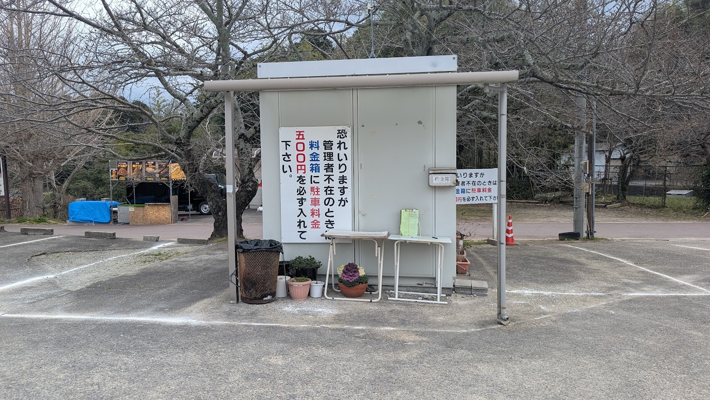信貴山朝護孫子寺の門前にある砂利敷き駐車場の料金箱。「管理人不在時はこちらへ」と書かれた立て看板と、代金を入れるための箱投入口。