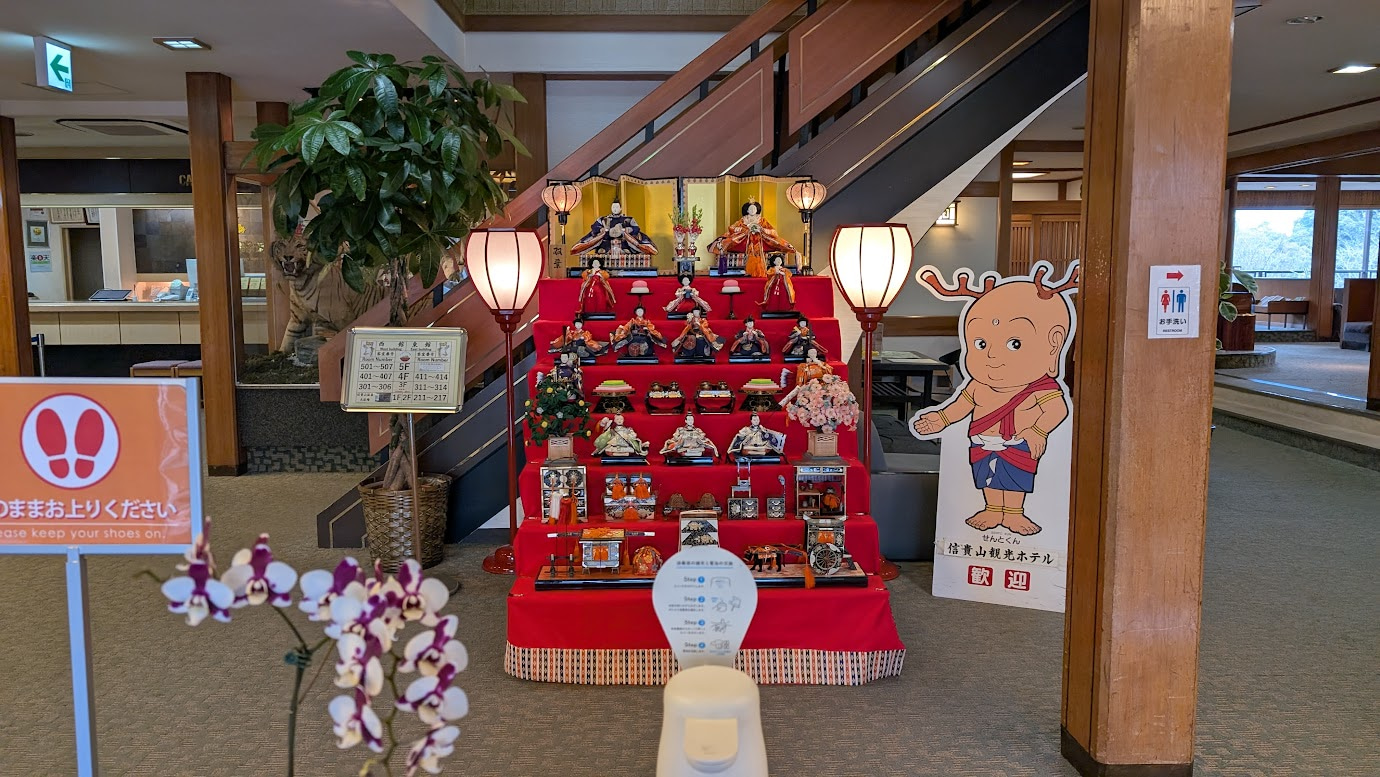 信貴山観光ホテルのロビーに飾られた豪華な七段飾りのひな人形。隣には奈良県の公式マスコットキャラクター「せんとくん」のパネルが立ち、宿泊客を歓迎している。