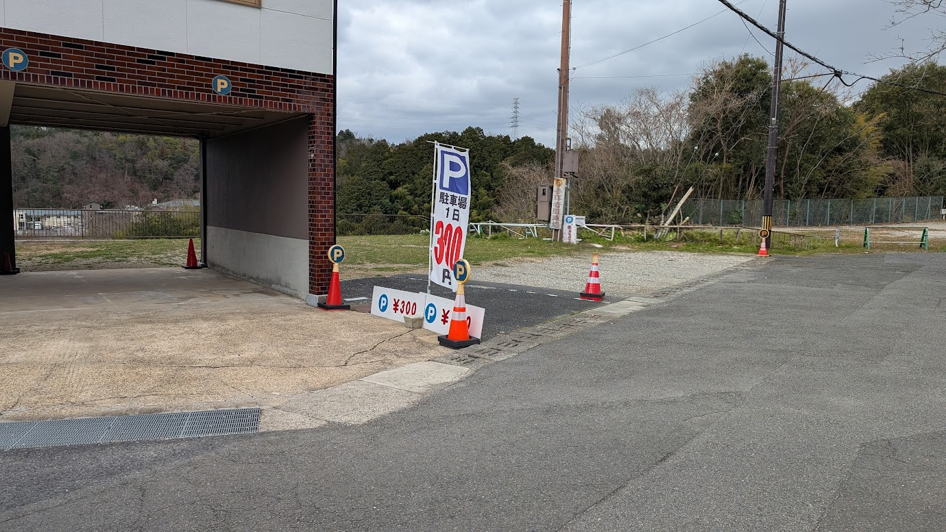 信貴山門バス停付近にある極小駐車場。手前は未舗装でロープ区画の300円スペース（5台）、奥には舗装された屋根付きの500円スペース（2台）が見える。