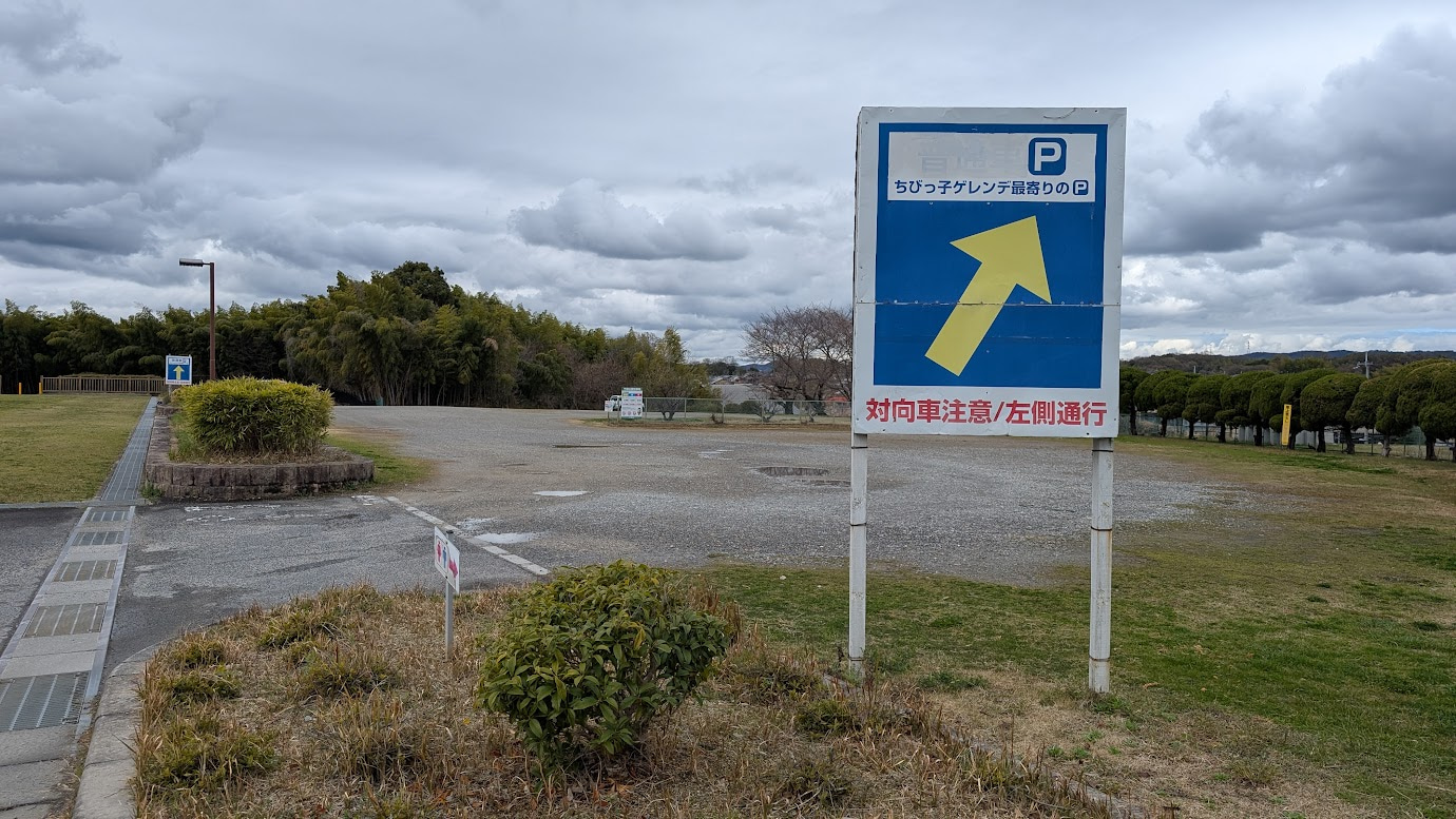 奈良県広陵町、竹取公園の奥にある広大な砂利敷きの駐車場。「ちびっこゲレンデ最寄りのP」と書かれた青い大きな看板があり、奥には数台の車が停まっている。