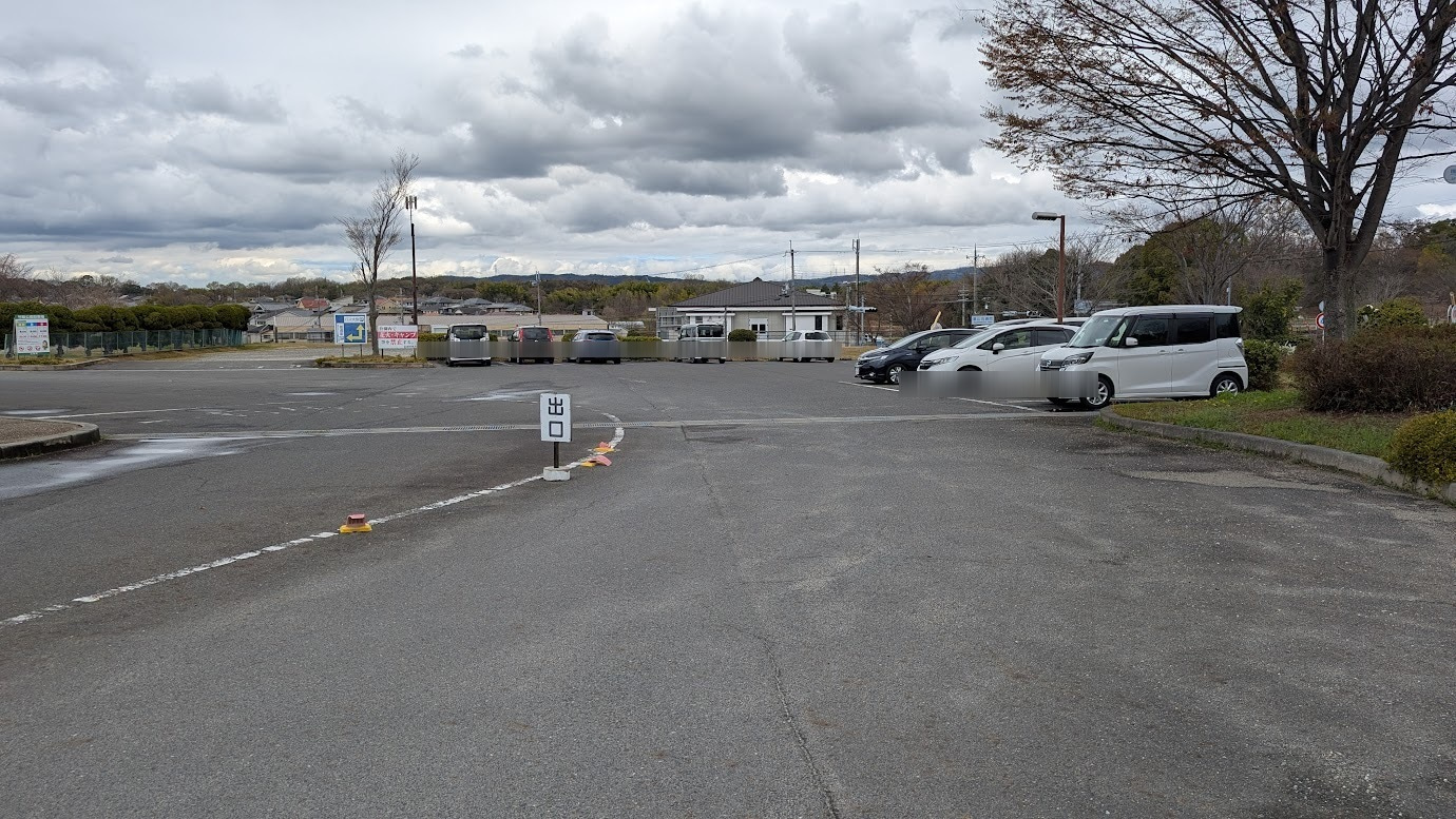 奈良県広陵町の竹取公園駐車場入口。平日の昼間、曇り空の下で多くの乗用車が駐車されており、入口付近は満車に近い混雑状況を示している。
