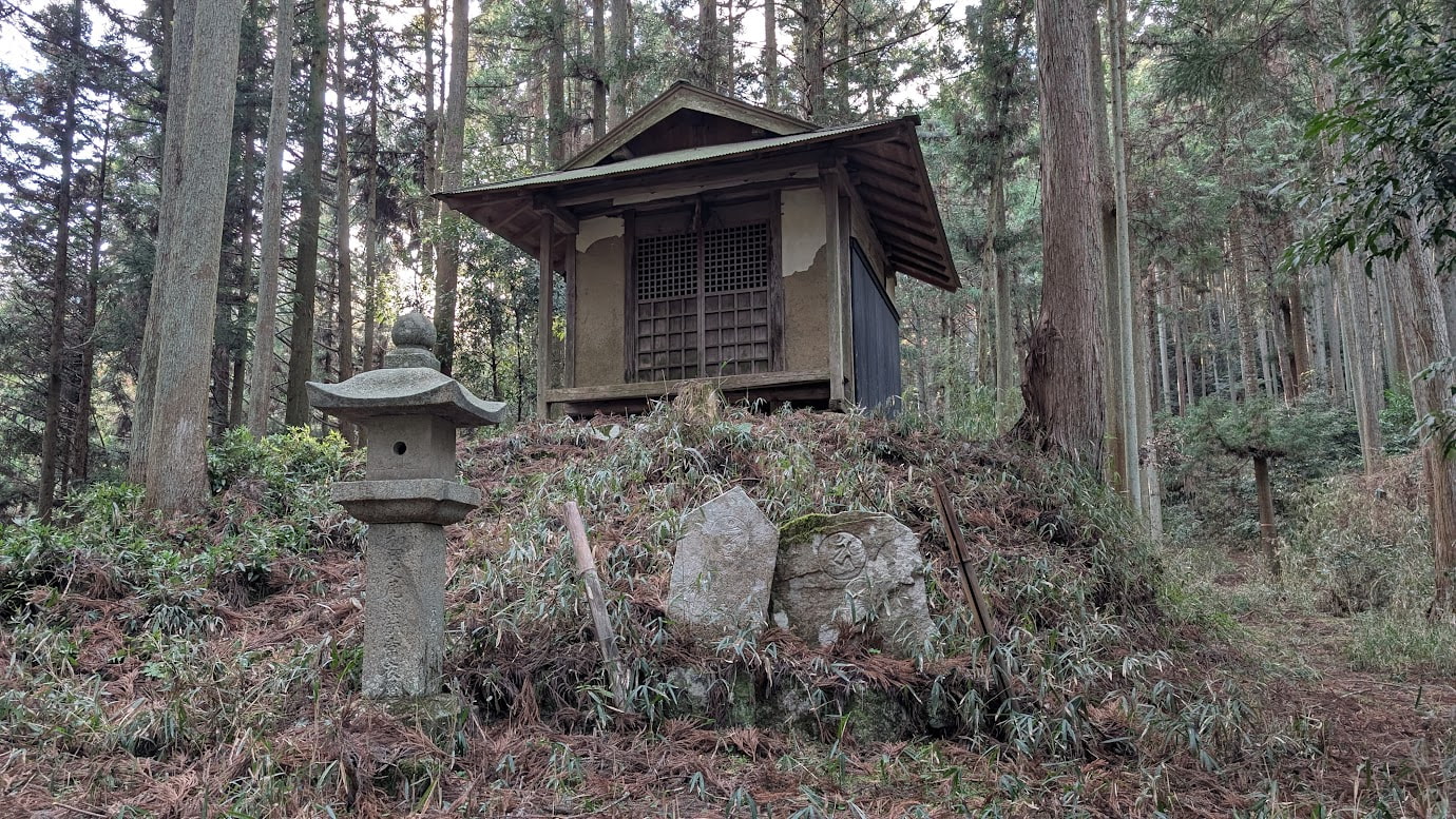 瀧蔵神社の薬師堂の全体。手前には歴史を感じる石灯籠と石碑が立ち、背後にはうっそうとした森が広がる静かな風景。
