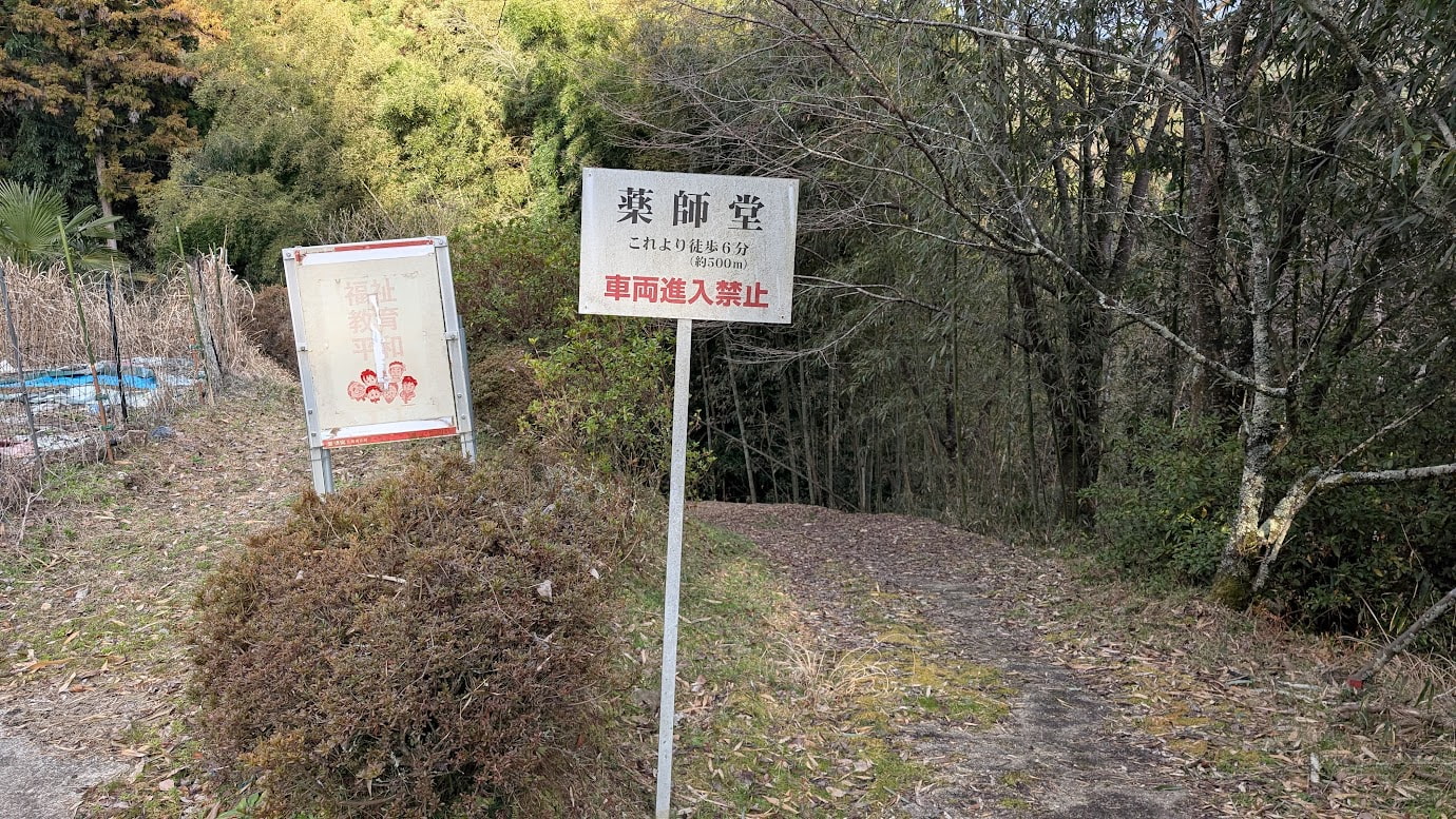 瀧蔵神社の駐車場下に設置された、木の案内板。手書きの日本語で「薬師堂への道」と「これより6分・約500m」と書かれている。