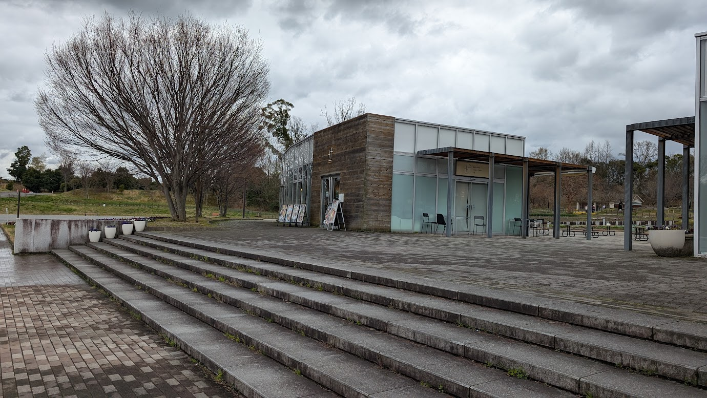 奈良県馬見丘陵公園内にある「Cafe. +f（カフェ・プリュススエフ）」の外観。木目調の壁と大きなガラス窓が特徴のモダンな平屋建てで、前には石造りの大階段とテラス席がある。