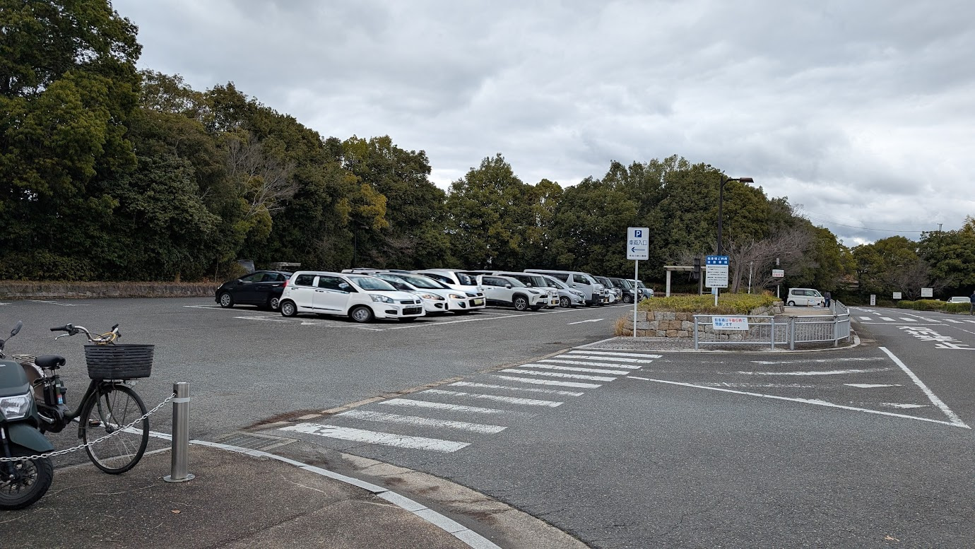 奈良県馬見丘陵公園の中央口駐車場。平日の昼間、曇り空の下で多くの車が駐車されており、満車に近い状態であることを示している。