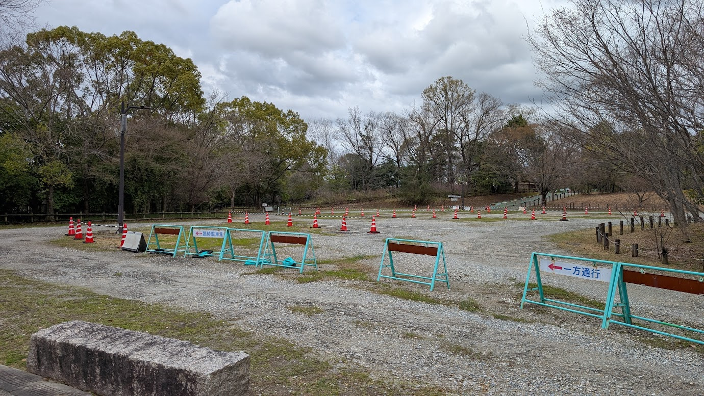 馬見丘陵公園の中央南口駐車場（臨時）。現在は閉鎖されており、広い駐車スペースには車がない。周囲には木々が立ち並び、遠くに公園の入り口ゲートが見える。