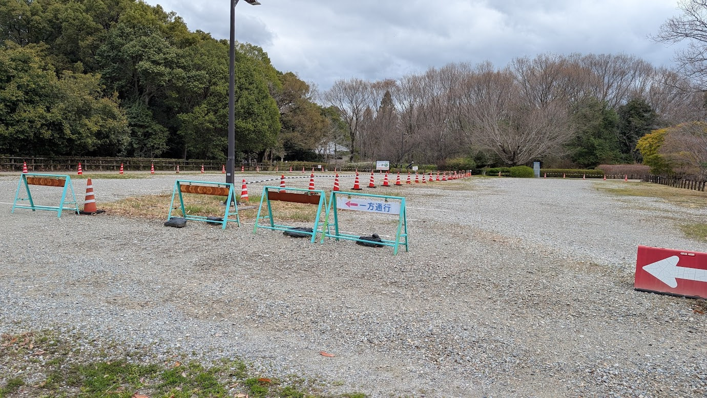 馬見丘陵公園の中央南口・上面駐車場。砂利敷きの広いスペースにカラーコーンや「一方通行」の案内が置かれている。背景には落葉した木々と深い緑の森が広がっている。