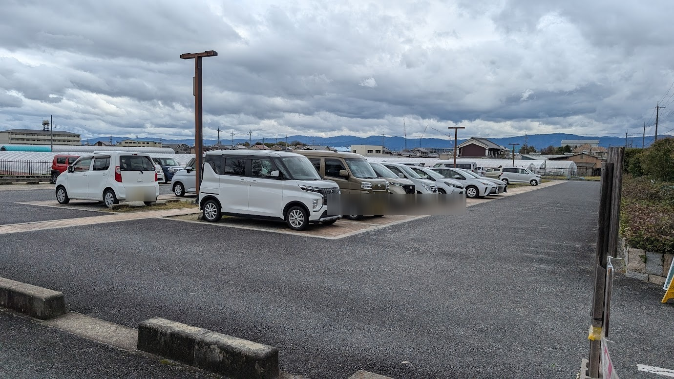 馬見丘陵公園の東駐車場。平日の昼間、曇り空の下で多くの乗用車が駐車されている。背景には遠くの山々とビニールハウスが広がる。