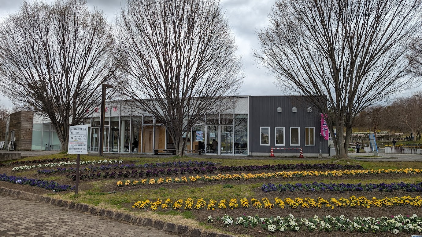 奈良県馬見丘陵公園の「花見茶屋」。手前には黄色、紫、白の花々が並ぶ花壇があり、奥には大きなガラス窓が特徴のモダンな休憩所が建っている。