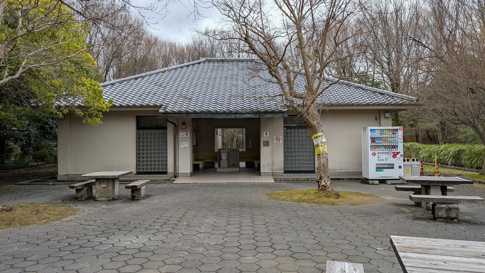馬見丘陵公園のナガレ山古墳近くにある公衆トイレの建物。瓦屋根の平屋建てで、手前には石畳の広場、石のベンチとテーブル、飲料の自動販売機が設置されている。