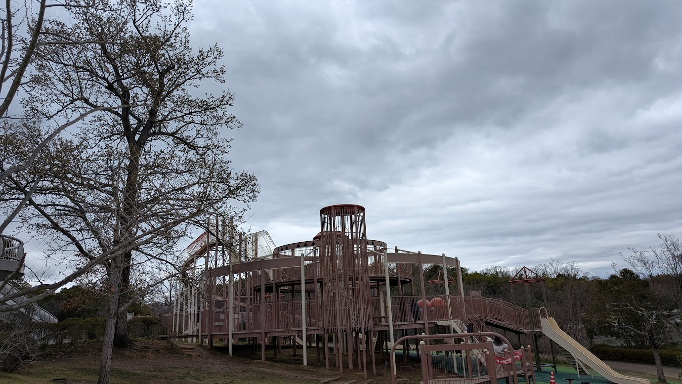 奈良県馬見丘陵公園の北エリアにある巨大な木造・金属製の大型遊具。スロープやタワー、長い滑り台が組み合わさっており、手前には葉の落ちた大きな樹木、背景には曇り空が広がっている。