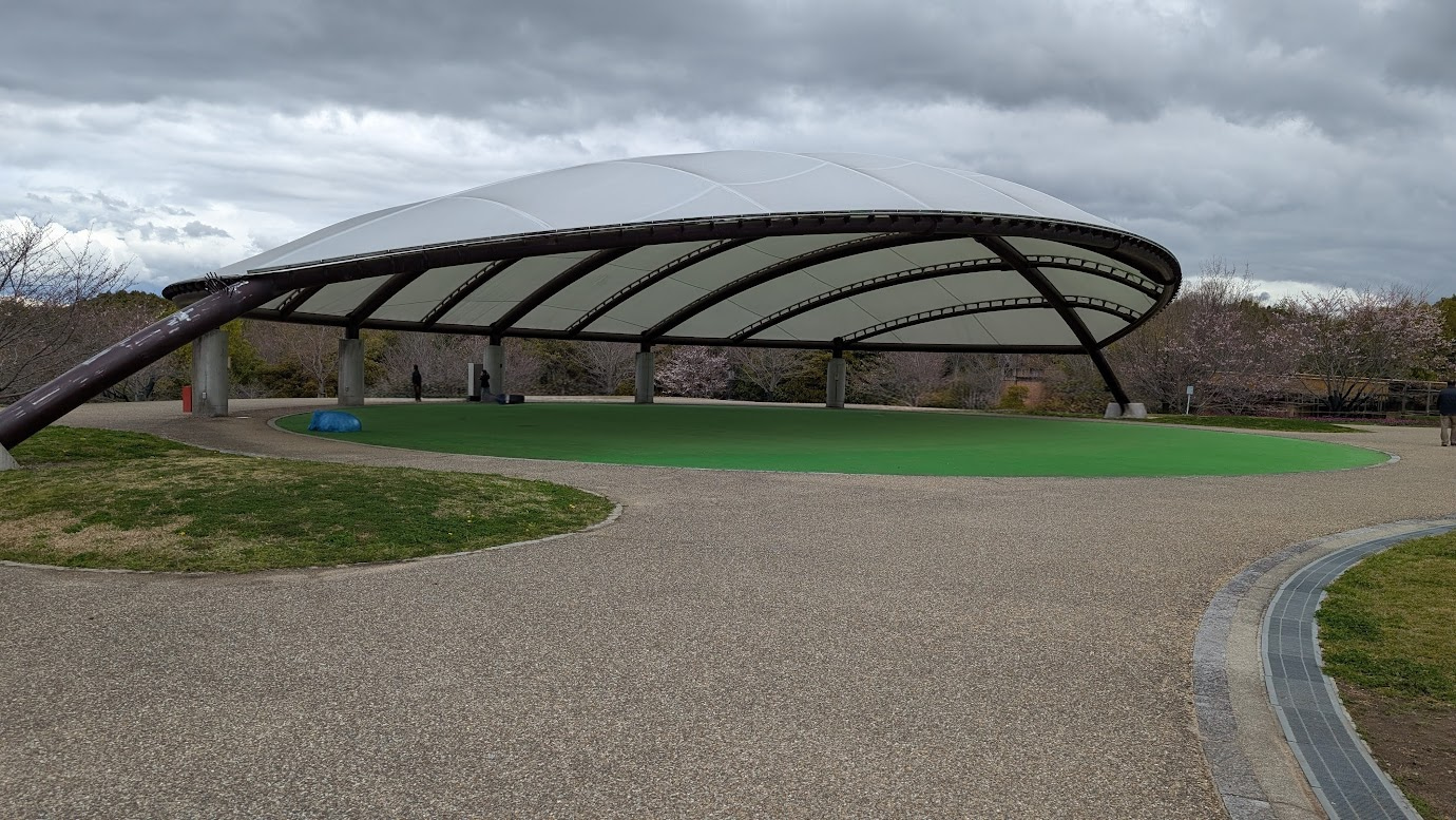 奈良県馬見丘陵公園の北エリアにある大型テント。白いドーム状の屋根の下に人工芝の緑の広場があり、周囲には咲き始めの桜の木々が並んでいる。