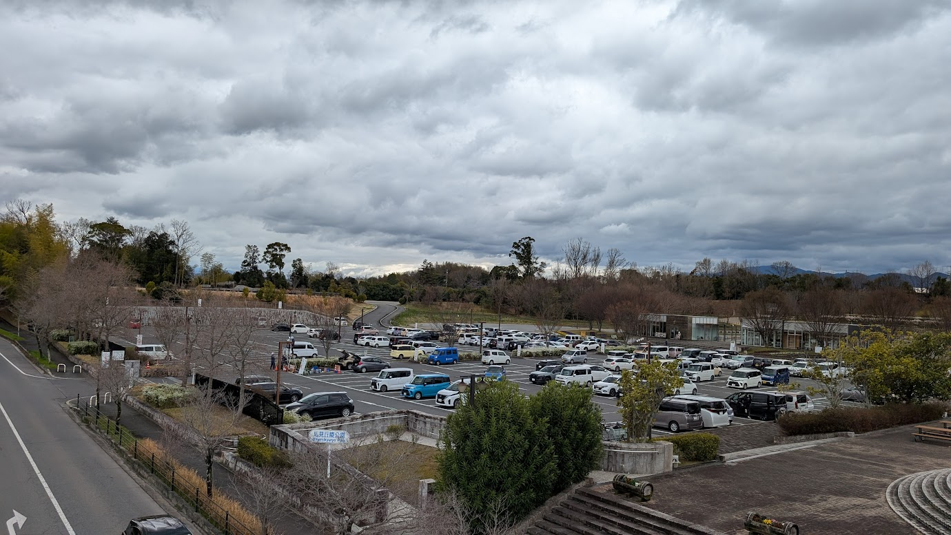 奈良県広陵町、馬見丘陵公園の北口駐車場の全景。平日の昼間、曇り空の下で多くの乗用車が駐車されている。手前には階段や植栽があり、奥には公園の入り口が見える。